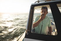 Um homem caucasiano aproveitando seu tempo de férias ao sol na costa, de pé em um barco, usando um walkie-talkie e falando — Fotografia de Stock
