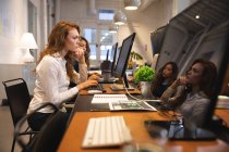 Кавказская предпринимательница, работающая в обычном современном офисе, сидящая за столом и пользующаяся компьютером с коллегой, работающим рядом с ней — стоковое фото