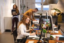 Business creativo femminile caucasico che lavora in un ufficio casual moderno, seduto a una scrivania e utilizzando un computer con colleghi che lavorano in background — Foto stock