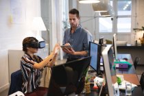 Misto razza femminile business creativo lavorando in un ufficio casual moderno, seduto a una scrivania e utilizzando una cuffia VR con un collega che lavora in background — Foto stock