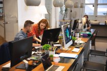 Кавказский женский и мужской бизнес-креативы, работающие в обычном современном офисе, сидящие за столом, за компьютером и разговаривающие с коллегой, работающим на заднем плане — стоковое фото