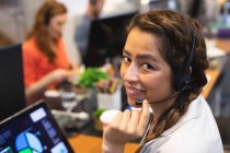 Porträt einer glücklichen Frau mit gemischter Rasse, die kreativ in einem lässigen, modernen Büro arbeitet, mit einem Kopfhörer telefoniert und in die Kamera schaut, während ihre Kollegen im Hintergrund arbeiten — Stockfoto