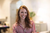 Портрет жінки-кавказької бізнес-творчості з довгим рудим волоссям, що працює в повсякденному сучасному офісі, посміхається і дивиться на камеру, одягнений в смугасту червону сорочку. — стокове фото