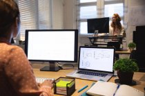 Au-dessus de la vue d'épaule d'une entreprise féminine mixte créative travaillant dans un bureau moderne décontracté, assis à un bureau et utilisant un ordinateur avec une collègue féminine travaillant en face d'elle au bureau debout — Photo de stock