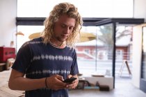 Kaukasischer Surfbrettmacher mit langen blonden Haaren, blauem T-Shirt und Holzschmuck steht in seinem Atelier und bedient sein Smartphone. — Stockfoto