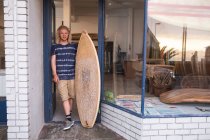 Homem caucasiano fabricante de pranchas de surf em pé na frente de seu estúdio, apoiando-se em uma moldura da porta da entrada, segurando uma prancha de surf novinha em folha e sorrindo . — Fotografia de Stock