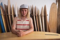 Retrato de un hombre caucásico fabricante de tablas de surf en su estudio, sosteniendo una de las tablas de surf y sonriendo a la cámara, con otras tablas de surf en un estante en el fondo. - foto de stock