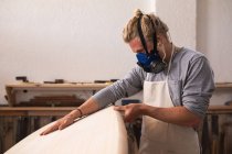 Kaukasischer Surfbrettmacher mit langen blonden Haaren, Gesichtsmaske, arbeitet in seinem Atelier, stellt ein Surfbrett her, inspiziert es und bereitet sich auf das Polieren vor.. — Stockfoto