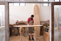 Kaukasischer Surfbrettmacher, der in seinem Atelier arbeitet, eine Schutzschürze trägt und sich eine Atemmaske aufsetzt, um ein Surfbrett zu polieren. — Stockfoto