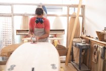 Kaukasischer Surfbrettmacher, der in seinem Atelier arbeitet, trägt eine Schutzschürze und eine Atemmaske und formt mit einem Schleifer ein hölzernes Surfbrett. — Stockfoto