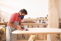 Homem caucasiano fabricante de pranchas de surf trabalhando em seu estúdio, vestindo um avental protetor e uma máscara de respiração, moldando uma prancha de madeira com uma lixadeira . — Fotografia de Stock