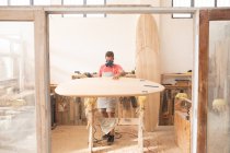Homme blanc fabricant de planches de surf travaillant dans son studio, portant un tablier de protection et un masque respiratoire, inspectant une planche de surf en bois lors de sa mise en forme avec une ponceuse. — Photo de stock