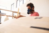 Homem caucasiano fabricante de pranchas de surf trabalhando em seu estúdio, vestindo um avental protetor e uma máscara facial de respiração, inspecionando uma prancha de madeira durante a modelagem com uma lixadeira . — Fotografia de Stock