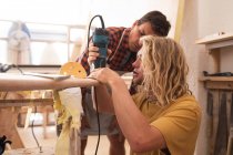 Dois fabricantes de pranchas de surf caucasianos trabalhando em seu estúdio e fazendo uma prancha de madeira juntos, polimento e moldando-a com uma lixadeira . — Fotografia de Stock