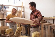 Dois fabricantes de pranchas de surf caucasianos trabalhando em seu estúdio e fazendo uma prancha de madeira juntos, inspecionando-a antes de pintar a superfície . — Fotografia de Stock