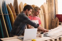 Zwei männliche Surfbrettmacher aus dem Kaukasus arbeiten in ihrem Studio, stehen am Türrahmen und arbeiten mit einem Laptop an Projekten, im Hintergrund Surfbretter — Stockfoto