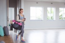 Caucásica atractiva bailarina de ballet con pelo rojo usando ropa deportiva, entrar en un estudio, preparándose para una clase de ballet, mirando su teléfono con auriculares en. - foto de stock