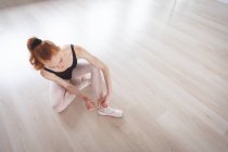 Bailarina de ballet caucásica atractiva con pelo rojo poniéndose sus zapatos de ballet, sentada en el suelo, preparándose para una clase de ballet en un estudio luminoso. - foto de stock