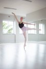 Dançarina de balé feminina caucasiana atraente com cabelo vermelho em pé em uma perna em sapatos pontiagudos durante a prática de balé em um estúdio brilhante, com foco em seu exercício com um braço levantado — Fotografia de Stock
