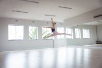 Кавказская привлекательная балетная танцовщица с рыжими волосами, танцующая балет, готовящаяся к занятию балетом в яркой студии, сосредоточенная на упражнениях, прыгающих в воздухе, с рукой над головой. — стоковое фото