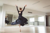Caucasian attractive female ballet dancer with red hair dancing ballet, wearing a black, long dress, preparing for a ballet class in a bright studio, focusing on her exercise. — Stock Photo