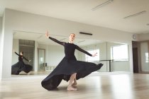 Ballerina di danza femminile attraente caucasica con balletto rosso per capelli, indossa un abito nero e lungo, si prepara per una lezione di balletto in uno studio luminoso, si concentra sul suo esercizio, sorridendo.. — Foto stock