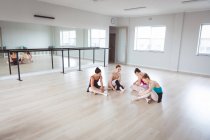 Um grupo de dançarinas de balé atraentes do sexo feminino caucasianas conversando em um estúdio de balé brilhante, parecendo felizes, se preparando para uma aula de balé, alongando e sentando no chão . — Fotografia de Stock