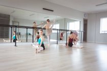 A group of Caucasian female attractive ballet dancers warming up, holding a barre and stretching on the floor in a bright ballet studio, focused on their exercise, preparing for a ballet class. — Stock Photo