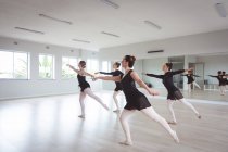 Un groupe de danseuses de ballet caucasiennes attirantes en tenues noires pratiquant pendant un cours de ballet dans un studio lumineux, dansant à l'unisson devant un miroir. — Photo de stock