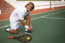 Portrait d'un homme caucasien portant des blancs de tennis passant du temps sur un court jouant au tennis par une journée ensoleillée, attachant des lacets de chaussures, regardant la caméra et souriant — Photo de stock