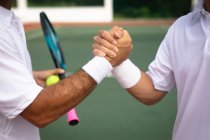 Seção intermediária fechar-se de homens vestindo brancos de tênis passar tempo em um campo juntos, jogando tênis em um dia ensolarado, apertando as mãos, um deles segurando uma raquete de tênis — Fotografia de Stock