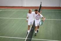 A Caucasian and a mixed race men wearing tennis whites spending time on a court together, playing tennis on a sunny day, embracing and smiling, holding tennis rackets — Stock Photo