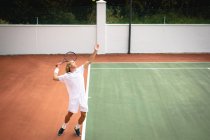 Ein kaukasischer Mann in weißen Tennisanzügen, der an einem sonnigen Tag auf einem Tennisplatz Tennis spielt, einen Tennisschläger in der Hand hält und sich darauf vorbereitet, einen Ball zu schlagen — Stockfoto