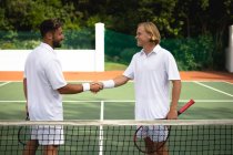 Um caucasiano e um misto vestindo brancos de tênis passando tempo em um campo juntos, jogando tênis em um dia ensolarado, apertando as mãos, um deles segurando uma raquetes de tênis — Fotografia de Stock