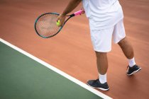 Sección media del hombre que usa blancos de tenis que pasan tiempo en una cancha jugando al tenis en un día soleado, sosteniendo una raqueta de tenis y preparándose para golpear una pelota - foto de stock