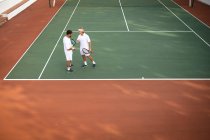Un caucásico y un hombre de raza mixta usando tenis blanco pasando tiempo en una cancha juntos, jugando al tenis en un día soleado, sosteniendo raquetas de tenis - foto de stock