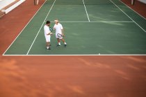 Ein kaukasischer und ein gemischter Rasse Männer in weißen Tennisanzügen verbringen Zeit auf einem Platz zusammen, spielen Tennis an einem sonnigen Tag und halten Tennisschläger in der Hand — Stockfoto