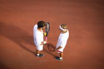 Um caucasiano e um misto vestindo brancos de tênis passando tempo em um campo juntos, jogando tênis em um dia ensolarado, segurando raquetes de tênis e uma bola — Fotografia de Stock