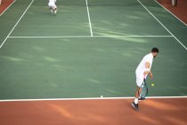 Ein kaukasischer und ein gemischter Rasse Männer in weißen Tennisanzügen verbringen Zeit auf einem Platz zusammen, Tennis spielen an einem sonnigen Tag, einer von ihnen bereitet sich auf einen Ball — Stockfoto