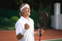 A Caucasian man wearing tennis whites spending time on a court playing tennis on a sunny day, holding a tennis racket, celebrating — Stock Photo