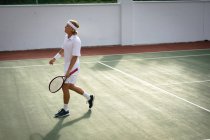 Un uomo caucasico che indossa i bianchi del tennis trascorre del tempo su un campo a giocare a tennis in una giornata di sole, con in mano una racchetta da tennis — Foto stock