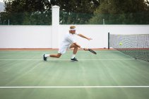 Un uomo caucasico che indossa i bianchi del tennis trascorre del tempo su un campo a giocare a tennis in una giornata di sole, tenendo in mano una racchetta da tennis, preparandosi a colpire una palla — Foto stock