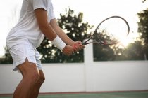 Середня секція чоловіка, який носить тенісні білі, проводить час на корті, граючи в теніс у сонячний день, тримаючи тенісну ракетку — стокове фото