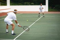 Ein kaukasischer und ein gemischter Rasse Männer in weißen Tennisanzügen verbringen Zeit auf einem Tennisplatz zusammen, spielen Tennis an einem sonnigen Tag, halten Tennisschläger und schlagen einen Ball — Stockfoto