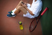 Средний вид на человека в теннисных белках, проводящего время на корте, играющего в теннис в солнечный день, сидящего на земле, используя смартфон, с теннисной ракеткой рядом — стоковое фото