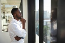 Un uomo d'affari afroamericano, indossa una camicia bianca, lavora in un ufficio moderno, guarda attraverso una finestra, tocca il mento e pensa: — Foto stock