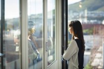 Uma mulher de negócios asiática trabalhando em um escritório moderno, olhando através de uma janela e pensando, cruzando os braços, em um dia ensolarado — Fotografia de Stock