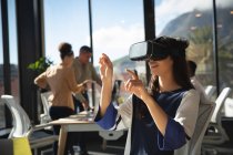 Une femme d'affaires asiatique travaillant dans un bureau moderne, portant un casque VR, touchant un écran virtuel interactif, avec ses collègues travaillant en arrière-plan — Photo de stock
