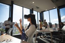 Une femme d'affaires asiatique travaillant dans un bureau moderne, portant un casque VR, touchant un écran virtuel interactif, avec ses collègues travaillant en arrière-plan — Photo de stock