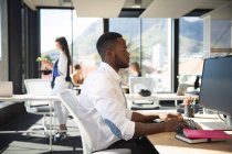 Ein afroamerikanischer Geschäftsmann, der in einem modernen Büro arbeitet, am Schreibtisch sitzt und einen Computer benutzt, während seine Kollegen im Hintergrund arbeiten — Stockfoto